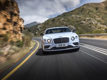 Een witte Bentley-auto met een Brits kenteken met de tekst "VER 45" rijdt op een bochtige bergweg, met een rotsachtige heuvel en groen gebladerte op de achtergrond, alsof hij de vraag van de AutoRAI WK-Quiz beantwoordt: waarin rijden de sterspelers?
