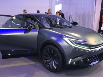 Binnen wordt een gestroomlijnde, futuristische Toyota bZ Compact SUV Concept met open bestuurdersdeur tentoongesteld. Verschillende mensen staan in de buurt en observeren het voertuig.