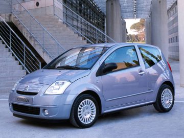 Een grijze Citroën C2 geparkeerd in een stedelijk gebied met metalen balustrades, trappen en architectonische constructies op de achtergrond.