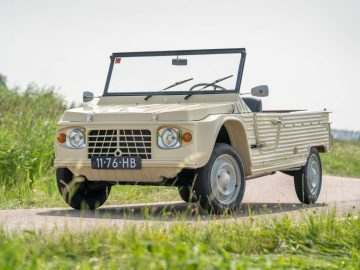 Een beige Citroën Mehari met cabriokap staat geparkeerd op een landweg, omgeven door weelderig groen.