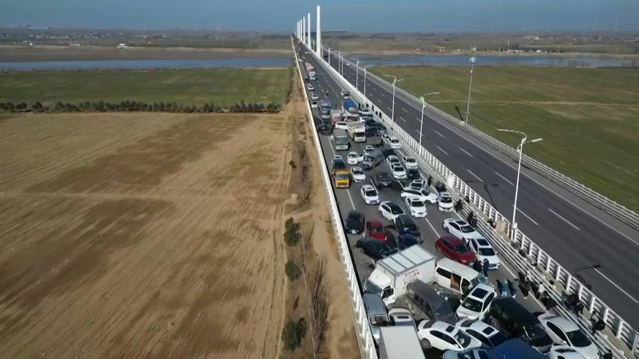 Luchtfoto van een snelweg met meerdere rijstroken met zwaar verkeer, inclusief auto's en vrachtwagens, die zich uitstrekt door een landelijk landschap met velden aan beide zijden. Temidden van de drukte blijkt er een kettingbotsing te zijn die voor flinke vertraging zorgt.