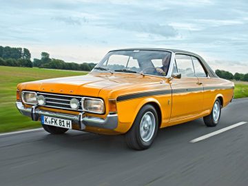 Een vintage oranje Ford Taunus rijdt op een weg met groene velden en bomen op de achtergrond onder een bewolkte hemel.