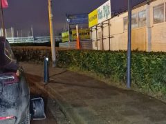 Een gedeeltelijk belemmerd trottoir naast een weg. Er staat een zwarte Tesla op het trottoir geparkeerd, waardoor de toegang voor voetgangers wordt belemmerd. Het gebied lijkt commercieel te zijn, met zichtbare borden en gebouwen.