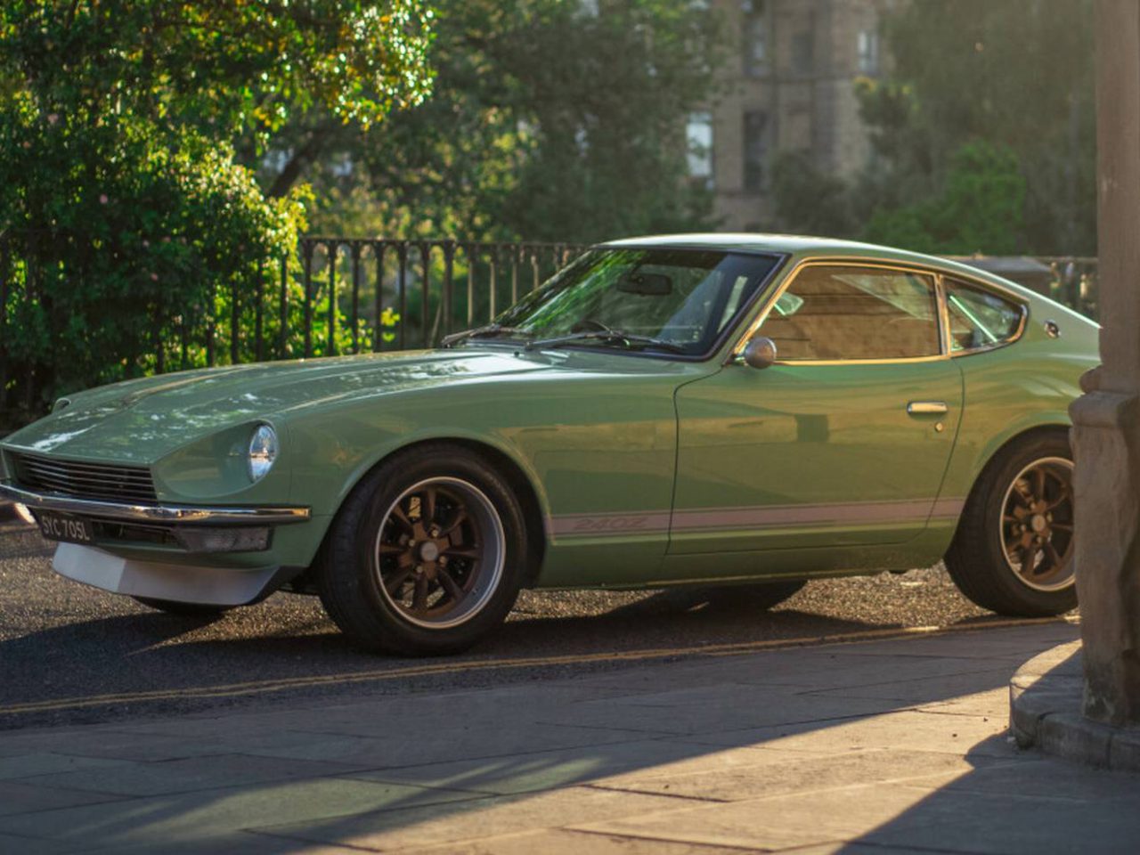 Een vintage groene Datsun 240Z staat geparkeerd in een stadsstraat, vlakbij een reling en wat groen, terwijl zonlicht schaduwen werpt.