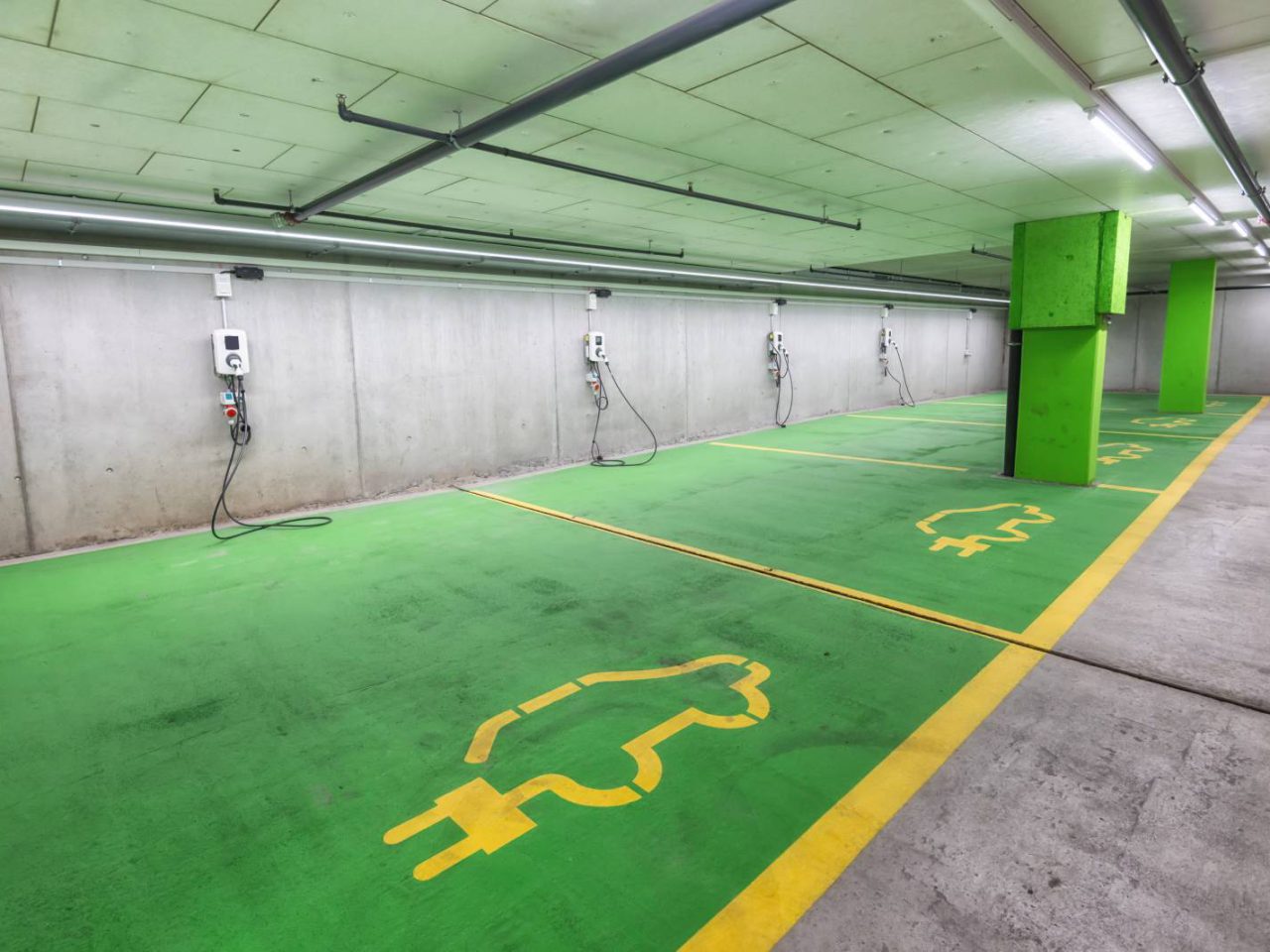 Leeg laadstation voor elektrische voertuigen binnenshuis met groen geschilderde vloeren, gele autosymbolen en aan de muur gemonteerde oplaadeenheden ontworpen voor elektrische auto's.