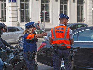 Twee politieagenten in uniform en hoge zichtbaarheidsvesten regelen het verkeer op een kruispunt. De ene agent wijst, terwijl de ander naast een zwarte auto staat. Er zijn een motorrijder en andere voertuigen zichtbaar.