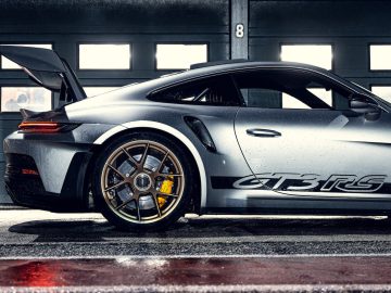Een zijaanzicht van een zilveren Porsche 911 GT3 RS-sportwagen met opgeheven achtervleugel, geparkeerd in een garage met het nummer 8 boven de deur weergegeven. De auto heeft strakke lijnen en gele remklauwen.