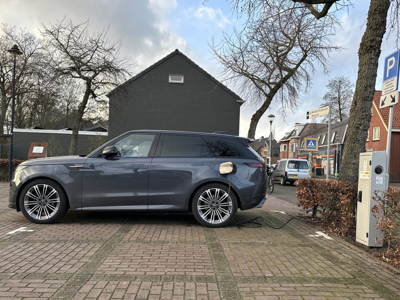 Een donkergekleurde elektrische Range Rover Sport staat geparkeerd op een daarvoor bestemde plek, aangesloten op een openbaar laadstation in een woonwijk. Bomen, huizen en een bewolkte lucht vormen de achtergrond in deze momentopname die elke Range Rover Sport-recensie waardig is.