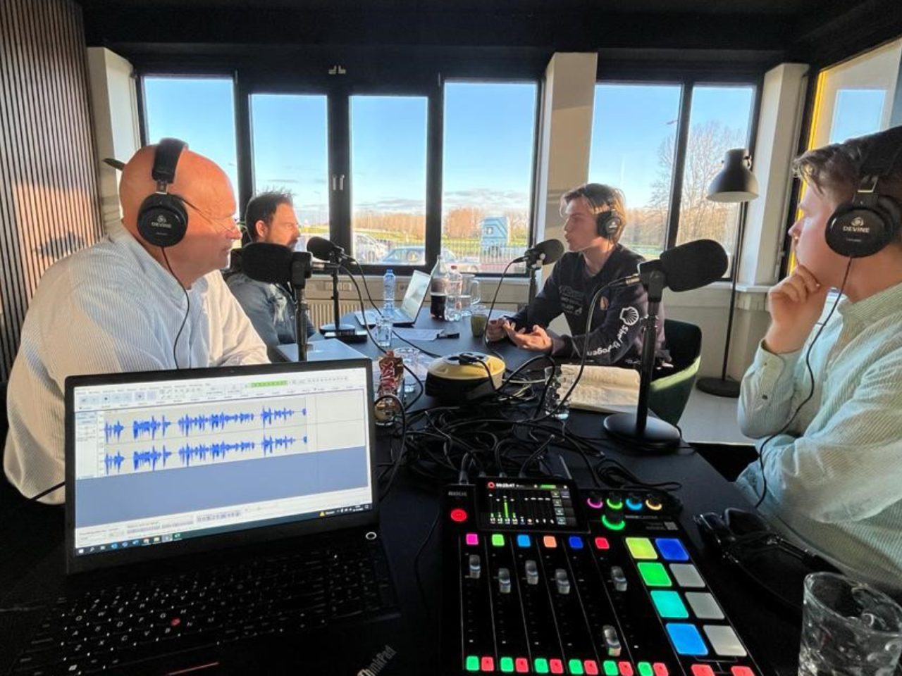 In een opnamestudio zitten vier mensen rond een tafel, elk met een koptelefoon op en in microfoons sprekend. Op de voorgrond zijn een laptop en een audiomixer met kleurrijke knoppen zichtbaar, die het decor vormen voor een boeiende AutoRAI Podcast-aflevering.