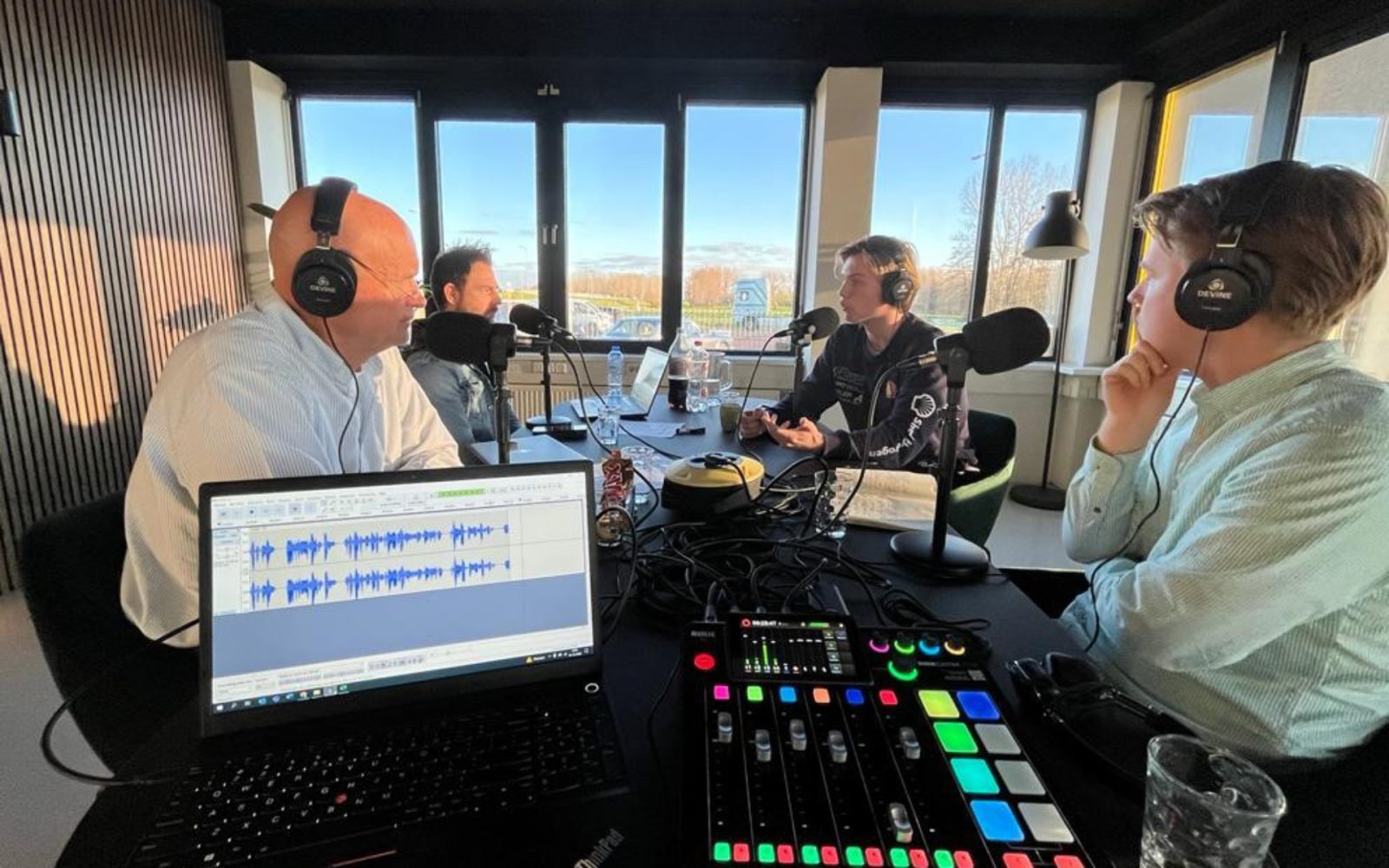 In een opnamestudio zitten vier mensen rond een tafel, elk met een koptelefoon op en in microfoons sprekend. Op de voorgrond zijn een laptop en een audiomixer met kleurrijke knoppen zichtbaar, die het decor vormen voor een boeiende AutoRAI Podcast-aflevering.