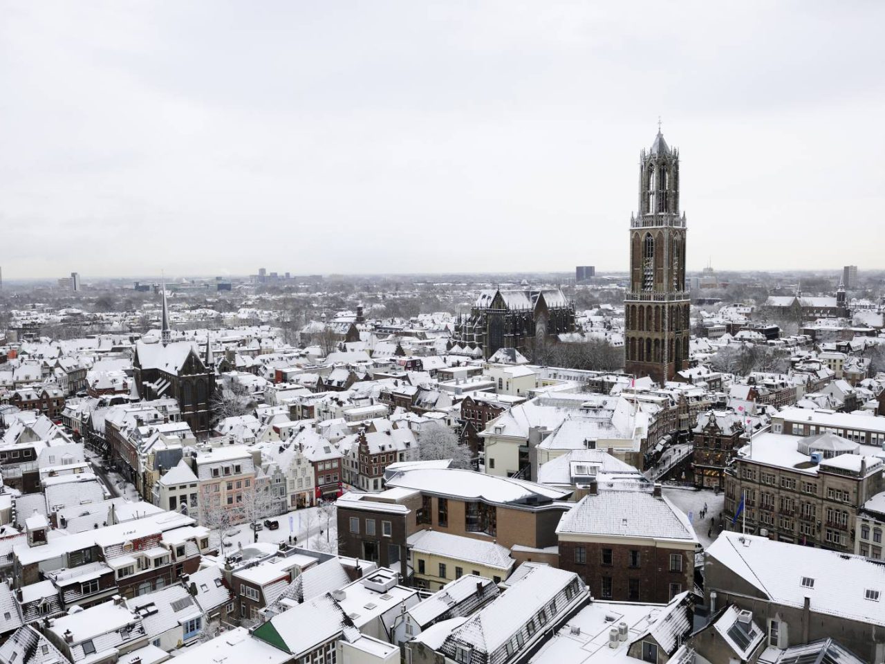 Luchtfoto van een met sneeuw bedekte stad, met een prominente hoge toren, talloze gebouwen met sneeuw op hun daken en voldoende plekken om gratis te parkeren.