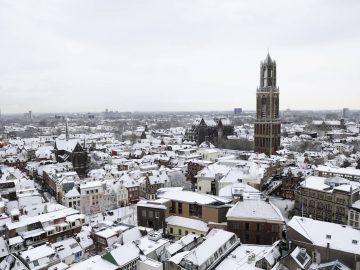 Luchtfoto van een met sneeuw bedekte stad, met een prominente hoge toren, talloze gebouwen met sneeuw op hun daken en voldoende plekken om gratis te parkeren.