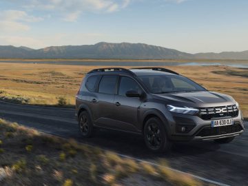 Een grijze Dacia Jogger Hybrid rijdt over een schilderachtige weg door een dor landschap met bergen op de achtergrond.