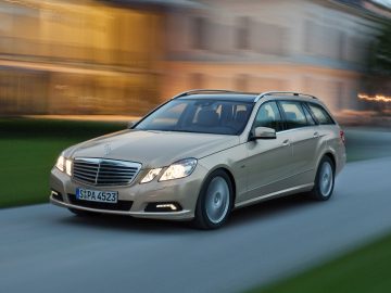 We zien een beige Mercedes-Benz stationwagen, een echte kilometervreter, rijden op een weg voor een vaag gebouw.