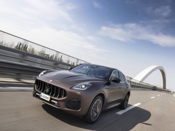 Een bruine Grecale SUV rijdt op een snelweg nabij een moderne boogbrug onder een gedeeltelijk bewolkte hemel.