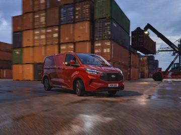 Een rood Ford Transit-busje rijdt door een scheepswerf beladen met gestapelde vrachtcontainers, met op de achtergrond een kraan tijdens de schemering.