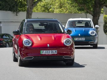 Twee compacte auto's rijden door een straat met bomen op de achtergrond. De voorste auto is een rode Ora Funky Cat met het kenteken "FJ22 DLN", en de auto erachter is blauw met het kenteken "FJ22 DLK".