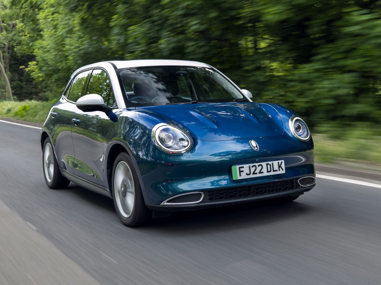 De Ora Funky Cat, een blauwe compacte auto met een wit dak, rijdt over een weg omgeven door groen. Op het kenteken staat "FJ22 DLK.