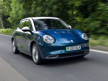 De Ora Funky Cat, een blauwe compacte auto met een wit dak, rijdt over een weg omgeven door groen. Op het kenteken staat "FJ22 DLK.