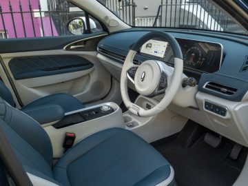 Een modern auto-interieur met tweekleurige blauwe en witte stoelen, een digitaal dashboarddisplay, een stuur met bedieningselementen en een open deur waardoor een roze buitenmuur en een zwarte reling aan de buitenkant zichtbaar zijn: stel je dit allemaal voor in de stijlvolle Ora Funky Cat.