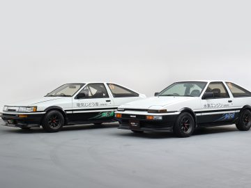 Twee witte Toyota Corolla AE86-auto's, naast elkaar weergegeven met Japanse tekst en afbeeldingen op de zijkant, geparkeerd op een grijze vloer met een effen achtergrond.