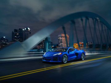 Een blauwe Corvette E-Ray-sportwagen rijdt 's nachts over een brug met de skyline van de stad en verlichte gebouwen op de achtergrond.