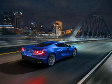 Een blauwe Corvette E-Ray rijdt 's nachts over een moderne brug, met een verlichte skyline van de stad op de achtergrond.