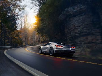 Een zilveren Corvette E-Ray rijdt bij zonsondergang over een kronkelige, met bomen omzoomde weg, met bladeren op de weg en een rotsformatie ernaast.