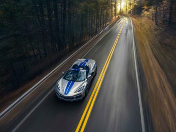 Een zilveren Corvette E-Ray met blauwe racestrepen rijdt op een bochtige weg door een bosrijke omgeving. De weg is omzoomd met bomen en de zon schijnt door het bos.