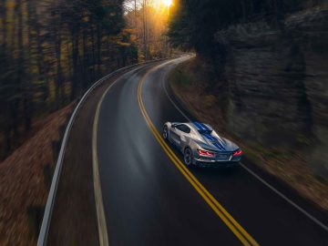 Een blauw-witte Corvette E-Ray rijdt over een kronkelige, met bomen omzoomde weg met herfstbladeren, richting zonlicht.