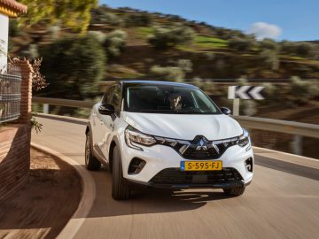 Een Mitsubishi ASX 2023 met Europees kenteken rijdt op een bochtige weg naast een bakstenen gebouw, bomen op de achtergrond.