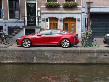 Een rode auto staat geparkeerd in een smal straatje aan een gracht, met fietsen en een groene slang in de buurt, terwijl een oplaadstation naast een gebouw staat met boogramen op de achtergrond.