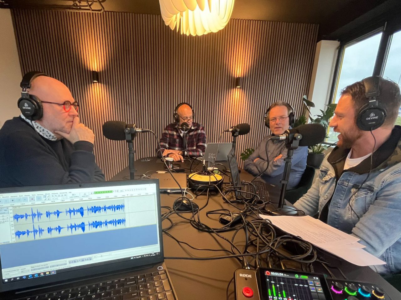 In een opnamestudio zitten vier mannen met microfoons rond een tafel, verwikkeld in de AutoRAI Podcast-discussie. Op de voorgrond is een laptop met audio-opnamesoftware zichtbaar.