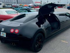 Een strakke zwarte limousine, precies vormgegeven als een Bugatti Veyron, staat majestueus tussen andere auto's buiten met de vleugeldeur open.