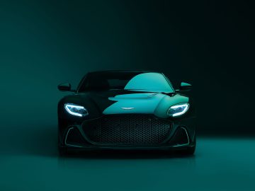 Vooraanzicht van een Aston Martin DBS 770 Ultimate, waarvan de donkergroene vorm elegantie uitstraalt met verlichte koplampen die door een donkere achtergrond met kleurverloop dringen.
