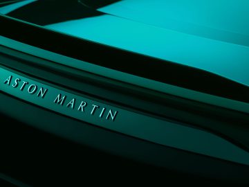 Close-up van de achterkant van een Aston Martin DBS 770 Ultimate, met de naam van het merk verlicht tegen het strakke, glanzende oppervlak onder groenblauw licht.