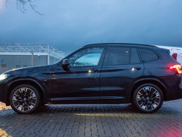 Zijaanzicht van een slanke zwarte BMW iX3 gefacelifte SUV, geparkeerd op een natte stenen stoep met een bewolkte hemel op de achtergrond.