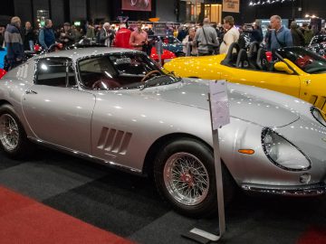 Een gepolijste zilveren vintage sportwagen met strakke rondingen en gedetailleerde kenmerken, te zien op Interclassics Maastricht, omringd door toeschouwers en andere kleurrijke auto's op de indoor autoshow.