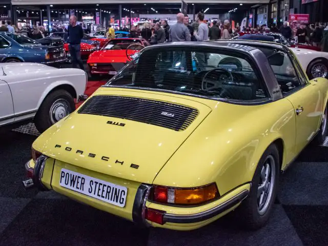 Een gele vintage Porsche met een kentekenplaat met de tekst "STUURBEKRACHTIGING" glinsterde onder de lichten van Interclassics Maastricht, binnenshuis tentoongesteld op een autoshow, omringd door andere klassieke auto's en enthousiaste bezoekers.