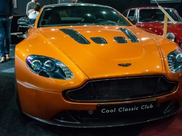 Een oranje sportwagen met zwarte grille en ventilatieopeningen in de motorkap wordt tentoongesteld in de Cool Classic Club tijdens het evenement Interclassics Maastricht. Op de achtergrond zijn andere voertuigen zichtbaar.