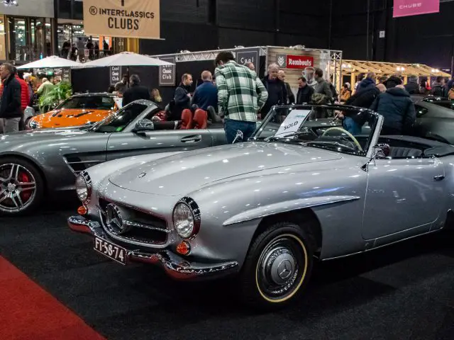 Op een autoshow wordt een verscheidenheid aan vintage en moderne auto's getoond, waaronder een zilveren cabriolet op de voorgrond met een bord 'Te koop' op de voorruit, dat doet denken aan de prachtige displays op Interclassics Maastricht.