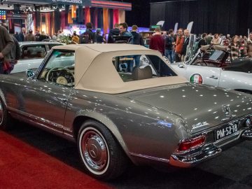 Op Interclassics Maastricht wordt een klassieke zilveren cabriolet met beige softtop tentoongesteld. Mensen staan eromheen en bewonderen verschillende tentoongestelde auto's.