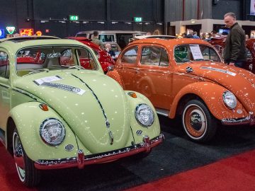 Op Interclassics Maastricht staan twee klassieke Volkswagen Kevers, één lichtgroen en één oranje, omringd door andere oldtimers en enthousiaste aanwezigen op de achtergrond.