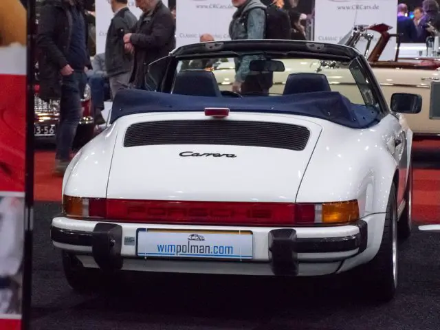 Op Interclassics Maastricht staat een witte Porsche Carrera cabriolet, van achteren gezien. Op het kenteken staat 'wimpolman.com'. Op de achtergrond zijn andere auto's en aanwezigen zichtbaar.