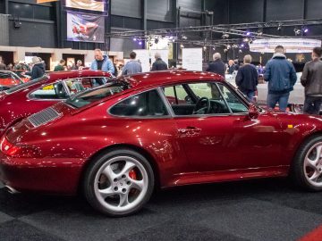 Een rode vintage sportwagen wordt tentoongesteld in een indoor autoshow op Interclassics Maastricht, terwijl mensen op de achtergrond andere voertuigen observeren.