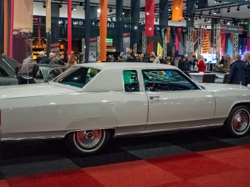 Een vintage witte coupé tentoongesteld op de indoor oldtimershow Interclassics Maastricht, met mensen en andere voertuigen op de achtergrond.