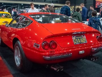 Op de indoor autoshow Interclassics Maastricht wordt een rode vintage sportwagen met kenteken "21X 029" tentoongesteld, omringd door mensen en andere klassieke auto's.