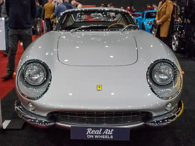 Een klassieke zilveren Ferrari-sportwagen tentoongesteld op de Interclassics Maastricht autoshow met een bord met de tekst "Real Art on Wheels", omringd door bezoekers en andere auto's.