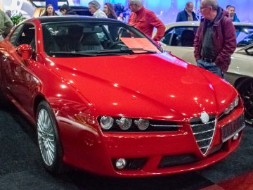 Op Interclassics Maastricht staat een rode sportwagen tentoongesteld, omringd door mensen en andere voertuigen op het prestigieuze indoor automotive evenement.