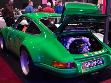 Op de indoor autoshow Interclassics Maastricht wordt een groene vintage sportwagen tentoongesteld met de motorruimte open. Verschillende mensen lopen rond en bekijken verschillende voertuigen.